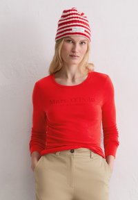 Rotes Langarmshirt mit dunkler Schrift, auf der "Marc O'Polo EST. IN STOCKHOLM" steht. Dazu eine gestreifte rot-weiße Mütze. Hosen in neutralen Farben.