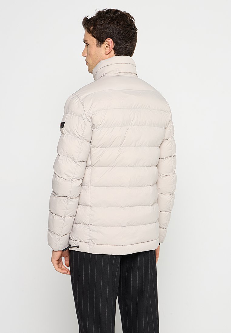 Giacca piumino beige con colletto alto, texture a cuciture orizzontali e tasche laterali con zip. Presenta un logo sulla spalla sinistra.