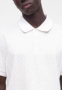 Homme portant un polo blanc avec un motif MK subtil répétitif et trois boutons, coupé au niveau du cou et du haut du torse.