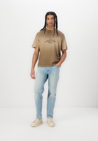 Brun bomulds T-shirt med grafisk tekst foran, parret med lyseblå slidte jeans og beige sneakers. Enkelt design med korte ærmer.