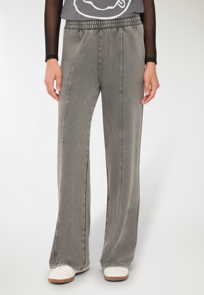 Osoba ubrana w luźne szare spodnie z przednimi szwami, białe sneakersy i czarną przezroczystą bluzkę z długim rękawem włożoną w pas.