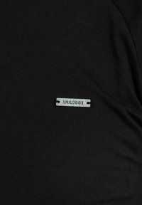 Metalen label met het woord "SMILODOX" op zwart ribbelstof genaaid, met zichtbare textuur en kleine plooien.