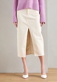 Falda de algodón crema con una abertura frontal. Combinada con un suéter de canalé morado claro y zapatos de tacón blanco. Diseño simple, hasta la rodilla.