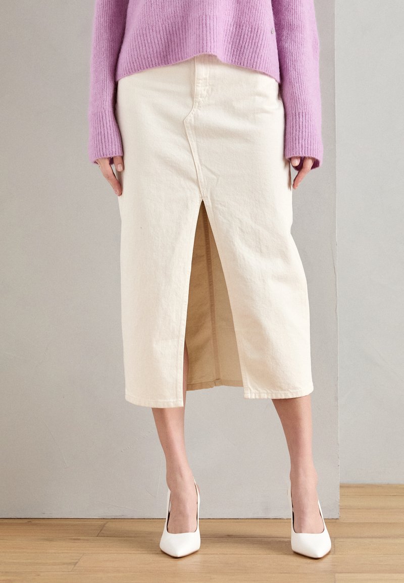 Falda de algodón crema con una abertura frontal. Combinada con un suéter de canalé morado claro y zapatos de tacón blanco. Diseño simple, hasta la rodilla.