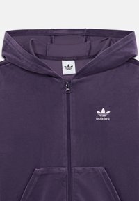 Lila bársonyos, cipzáros kapucnis pulóver, elülső zsebekkel és egy kis fehér Adidas logóval a bal mellen. Sima textúra, laza dizájn.