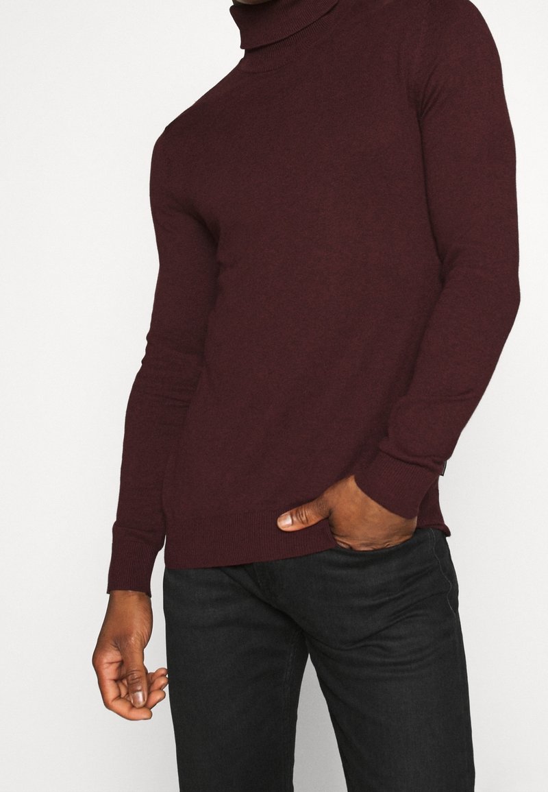 Pull à col roulé bordeaux en tissu doux, avec manches longues et poignets côtelés. Porté avec un jean foncé sur un fond uni.