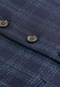 Gros plan d'un tissu à carreaux bleu foncé avec deux boutons noirs étiquetés "Baker" cousus dessus.