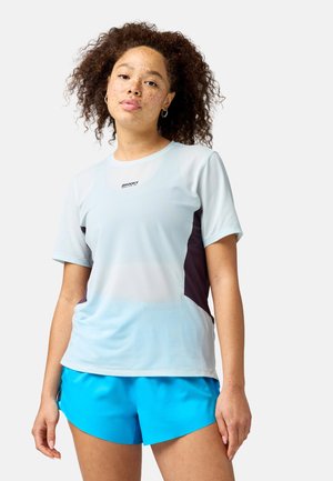 Jeune femme avec des taches de rousseur, cheveux bouclés, portant un t-shirt Brooks gris clair avec des panneaux latéraux noirs et un short de sport bleu vif.
