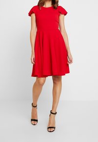 Czerwona sukienka z krótkim rękawem, dopasowanym gorsetem, rozkloszowaną spódnicą i falbanami na rękawach. Noszona z czarnymi szpilkami, ustawiona na neutralnym tle.