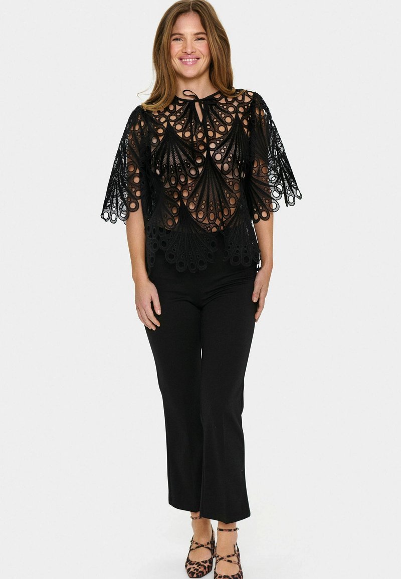 Top in pizzo nero con motivi intricati e maniche a campana, dotato di un collo con lacci. Abbinato a pantaloni neri e scarpe con stampa animale.