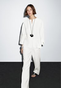 Vrouw met kort bruin haar, gekleed in een losse witte geknoopte top en wijde witte broek, een zwarte hangerketting, een zilveren armband en zwarte sandalen.