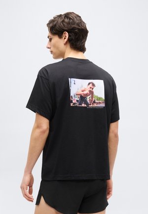 Młody mężczyzna widziany od tyłu, ubrany w czarny T-shirt z nadrukiem osoby biegnącej oraz napisem „Biegacze, jesteśmy inni” na plecach.