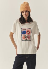 Biała bawełniana koszulka z kolorowym wzorem stempla kwiatowego z niebieskimi, różowymi i pomarańczowymi kwiatami. Do tego pomarańczowa czapka dzianinowa o fakturowanej strukturze.