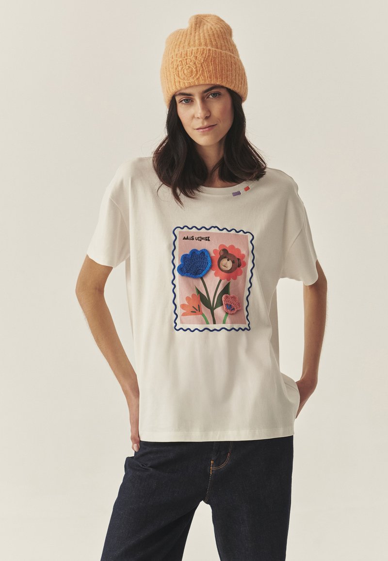 Biała bawełniana koszulka z kolorowym wzorem stempla kwiatowego z niebieskimi, różowymi i pomarańczowymi kwiatami. Do tego pomarańczowa czapka dzianinowa o fakturowanej strukturze.