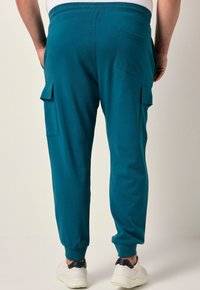 Teal bomullsbyxor med ribbade fållar, cargofickor på sidorna och en bakficka, i kombination med vita sportskor.