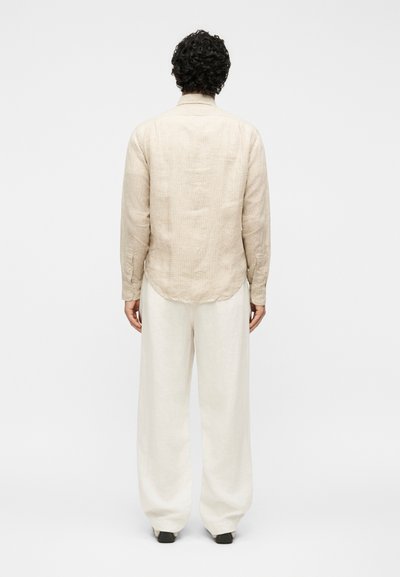 Άτομο με σγουρά σκούρα μαλλιά, που φαίνεται από πίσω, φοράει μπεζ μακρυμάνικο πουκάμισο και φαρδιά άσπρα παντελόνια σε απλό φόντο.