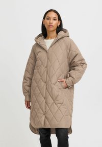 Manteau matelassé beige avec capuche, doté d'une fermeture à boutons, de poches latérales et d'une coupe longue et décontractée. Motif en losanges texturé sur l'ensemble.