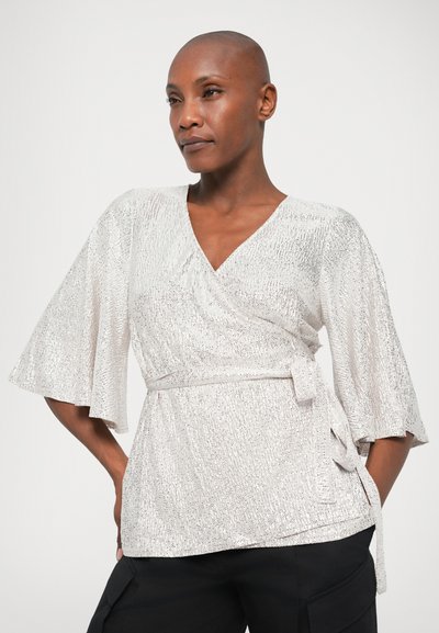 Metallisk silverfärgad wrap-top med korta utsvängda ärmar, texturerad yta och detalj med knytmidja. Bärs över mörka byxor.