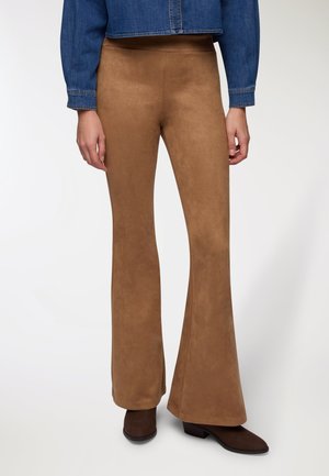 Donna che indossa pantaloni a zampa d'elefante in suede marrone, stivali con tacco marroni scuri e una giacca di jeans blu cropped, in piedi contro uno sfondo semplice.