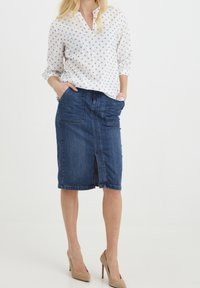 Gonna a tubino in denim con spacco frontale e molteplici tasche. Abbinata a una blusa leggera bianca a maniche lunghe con piccoli motivi floreali blu.