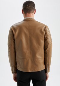 Uomo con una giacca in pelle color beige aderente, con maniche trapuntate e polsini con zip, in piedi di spalle su uno sfondo bianco uniforme.