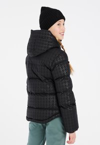 Giubbotto piumino nero con motivo a scacchi, cappuccio aderente, chiusura con zip e una texture elegante e trapuntata. Abbinato a un berretto in maglia nera.