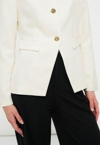 Blazer blanc cintré avec deux boutons décoratifs dorés et poches à rabat, assorti à un pantalon noir.