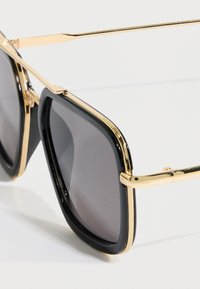 Μαύρα τετράγωνα γυαλιά ηλίου με γυαλιστερό φινίρισμα, διακοσμημένα με χρυσές μεταλλικές λεπτομέρειες και σκούρους φακούς. Ο σκελετός έχει τολμηρό, μοντέρνο σχεδιασμό.