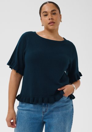 Nő sötétkék rövid ujjú kötött felsőben fodros szegéllyel és világoskék farmerral, egyik kezével a zsebében pózolva fehér háttér előtt.