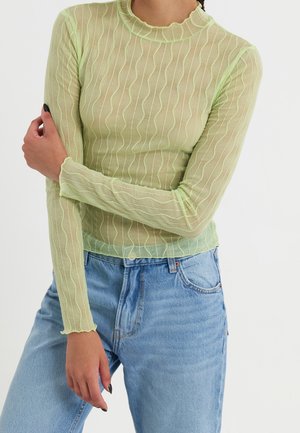 Femme portant un haut transparent vert citron à manches longues avec des rayures ondulées et un jean taille haute bleu clair sur fond blanc.