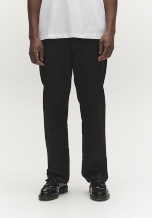 Mees, kes kannab musti sirglõikelisi pükse, valget särki ja musti nahast sassidega loafereid, seisab tavalise hele tausta ees.