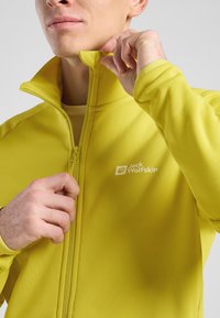 Gele softshell jas met hoge kraag, ritsluiting en een logo op de linkerborst. Glad textuur en een aansluitend ontwerp.