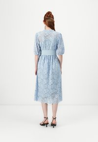 Lyseblå blond midi-kjole med korte pufferm, innsvingt midje og scallopkant, som viser intrikate blomster mønstre.