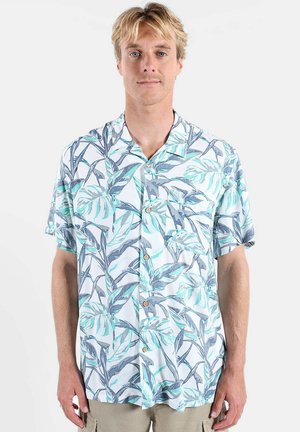 Homme aux cheveux blonds portant une chemise blanche à manches courtes avec des motifs de feuilles bleues et vertes, debout devant un fond uni.
