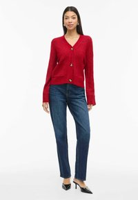 Žena u crvenom kardiganu sa gumbima i prednjim džepovima, plavim trapericama i crnim cipelama s oštrim vrhom, stoji pored bijele pozadine.