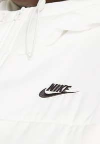 White hooded jacket made of smooth fabric, featuring a black embroidered Nike logo on the chest, with adjustable drawstring and zipper.