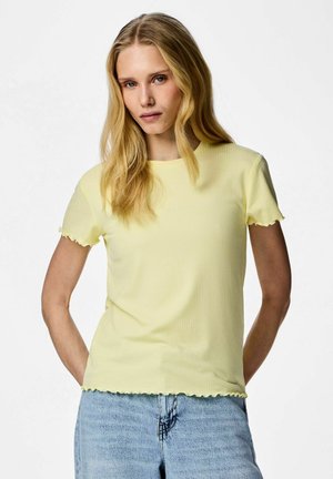 Mujer rubia con una camiseta de manga corta amarillo claro con borde ondulado y jeans de mezclilla azul claro, de pie con las manos detrás de la espalda.