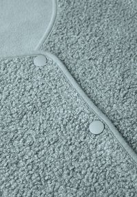 Tessuto grigio texture con finitura riccia, caratterizzato da bordi lisci e bottoni a pressione bianchi lungo la cucitura. Aspetto morbido e invitante.