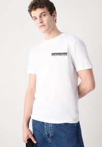 Balta medvilninė T-maišelis su juodu stačiakampiu logotipu, kuriame yra užrašas "Calvin Klein New York" kairiajame krūtinės šone. Paprastas trumpomis rankovėmis dizainas.
