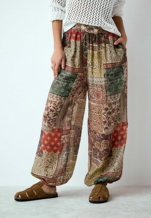 Személy laza, mintás foltvarrott nadrágot visel rugalmas passzéval, fehér kötött pulóvert és barna, lábujjakat szabadon hagyó szandált, zsebre tett kézzel áll.