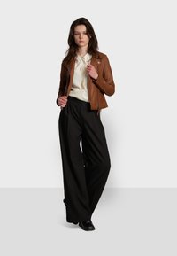 Giacca di pelle marrone con chiusura a zip sopra una camicetta bianca, abbinata a pantaloni larghi neri e stivaletti neri. Design semplice.