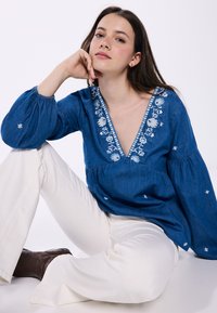 Blaue Denim-Bluse mit Puffärmel, V-Ausschnitt und weißer Blumenstickerei. Kombiniert mit weißen Hosen und braunen Stiefeln.