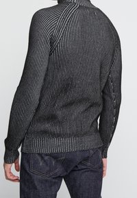 Svart och grå ribbad tröja med långa ärmar, som har en hög krage och ett strukturerat stickat mönster, parat med mörka denimjeans.