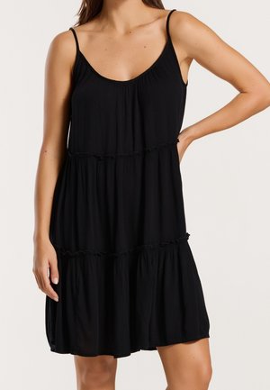 Kobieta w czarnej, warstwowej sukience bez rękawów na cienkich ramiączkach, stojąca z jedną ręką zgiętą i opartą na biodrze na jednolitym tle.