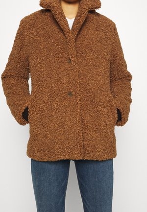 Brun texturerad ullkappa med krage, knäppning fram och sidofickor. Den är kombinerad med blå denimjeans och har en lurvig yta.