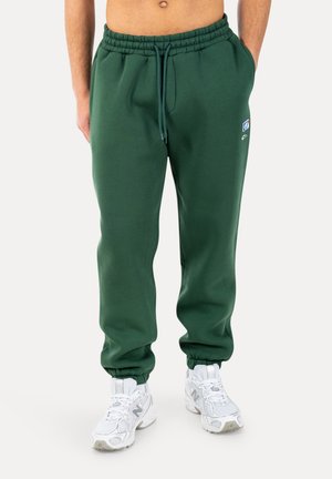 Uomo che indossa pantaloni da jogging verde scuro con vita elastica e coulisse, e sneakers bianche New Balance, in piedi con le mani nelle tasche.