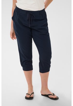 Femeie purtând pantaloni scurți bleumarin cu șnur și șlapi negri, stând pe un fundal alb.