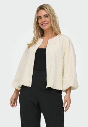 Frau lächelnd, trägt eine off-white strukturierte Reißverschlussjacke mit weiten Ärmeln über einem schwarzen, mit Spitze besetzten Top und schwarzen Hosen.
