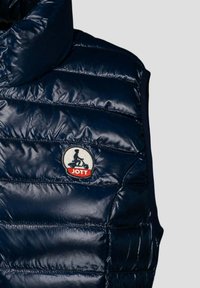 Navyblauer Steppwesten mit Stehkragen, gestepptem Muster und einem runden JOTT-Logo-Patch mit einer Motorrad-Silhouette. Glänzende Textur.