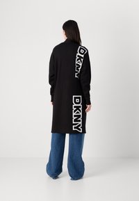 Črn dolg plašč z drzno belim napisom "DKNY" na hrbtni strani, v kombinaciji s širokimi jeansi. Gladka tekstura in sproščen kroj.